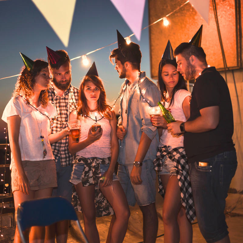 A group of young adults gathered outdoors during the evening, wearing party hats and surrounded by string lights, thinking and engaged in quiet conversation.