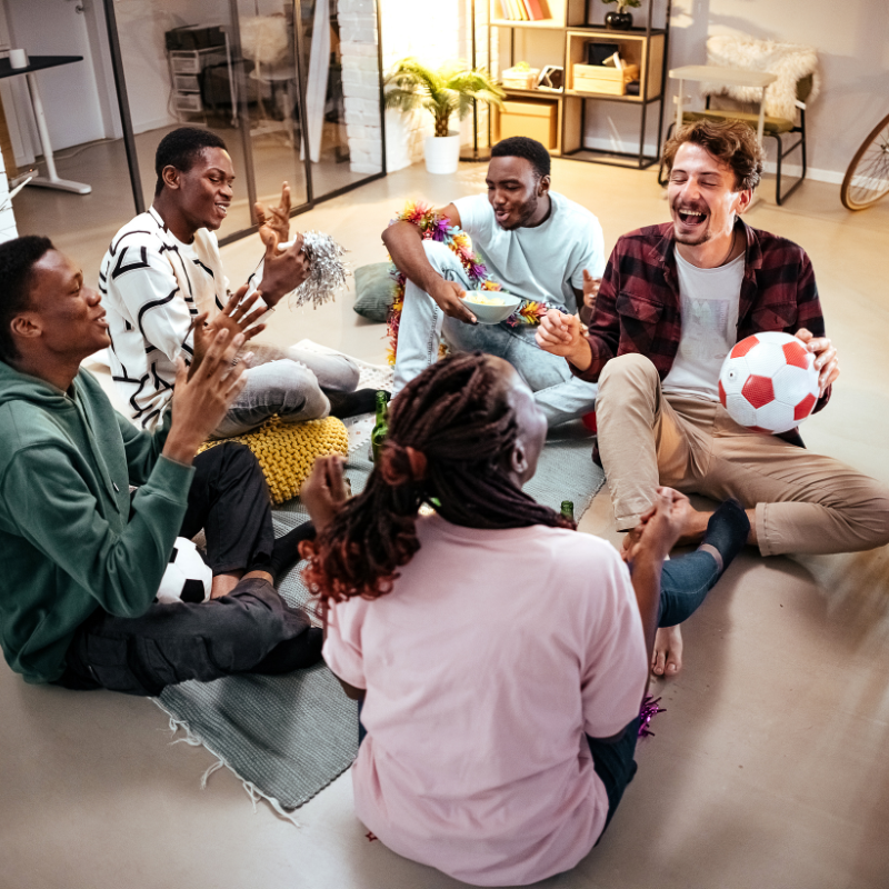 A group of five friends sitting on the floor in a cozy living room, laughing and enjoying a casual gathering. One holds a soccer ball, while others engage in animated conversation.