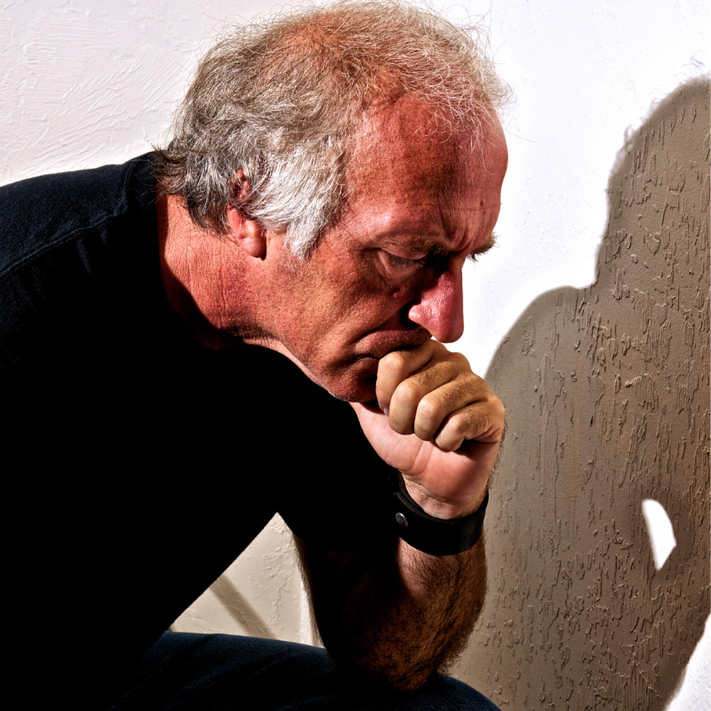 An older man deep in thought, resting his chin on his hand, with a solemn expression and a shadow cast on the wall behind him, reflecting the weight of pain.