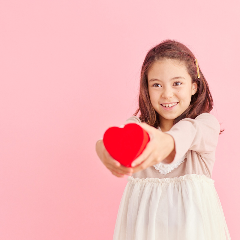 A smiling young girl with dark hair joyfully extends a red heart-shaped box toward the viewer, symbolizing the open and generous flow of love. The soft pink background enhances the warmth and kindness of the moment.