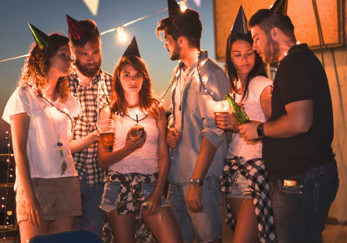 A group of young adults gathered outdoors during the evening, wearing party hats and surrounded by string lights, thinking and engaged in quiet conversation.