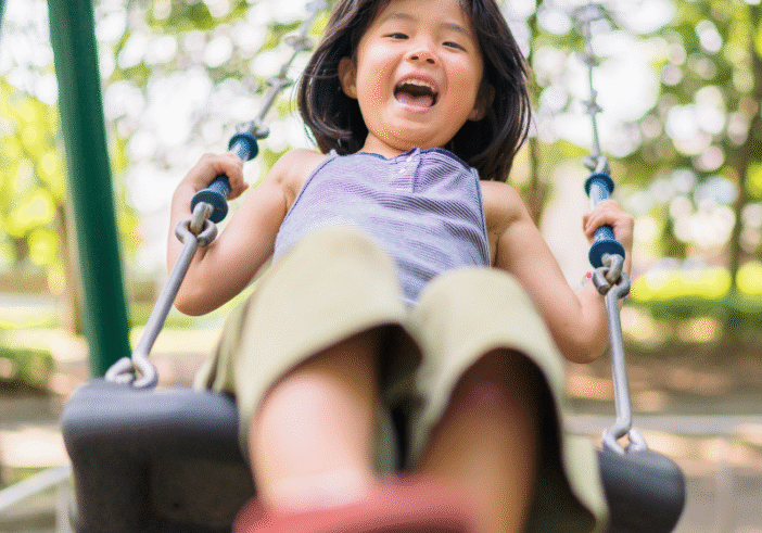 A young girl laughing joyfully while swinging in a sunny park, radiating child-like aliveness and energy.