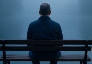 Middle-aged man sitting alone on a bench facing a foggy blue lake
