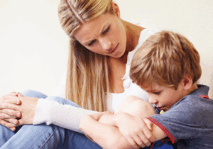 A young boy sitting with his knees pulled to his chest, showing visible emotion, while a concerned woman gently listens and offers support.