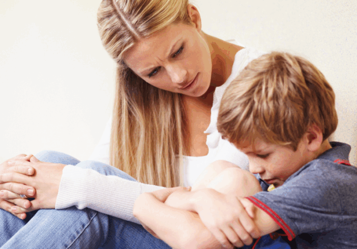 A young boy sitting with his knees pulled to his chest, showing visible emotion, while a concerned woman gently listens and offers support.