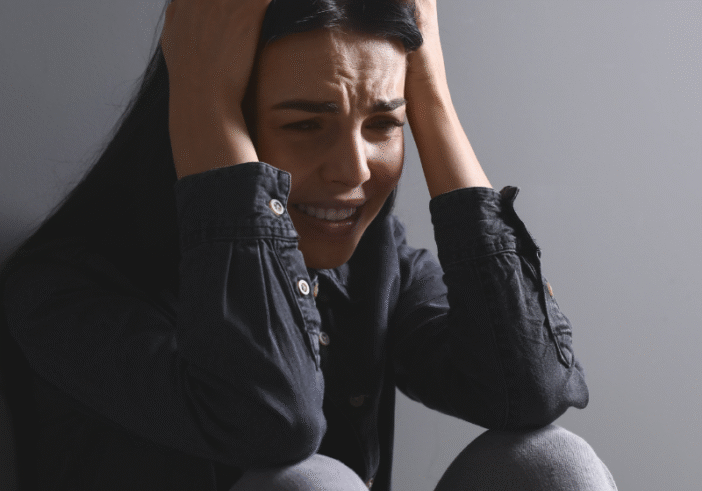 A distressed woman sitting against a gray wall, holding her head in her hands, reflecting feelings of uncertainty and worry about the year 2025.